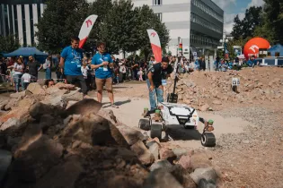 Robot poruszający się po terenie z kamieniami, z uczestnikami wydarzenia w tle.