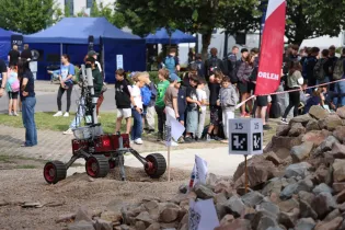 Robot poruszający się po terenie z grupą ludzi w tle i namiotami.