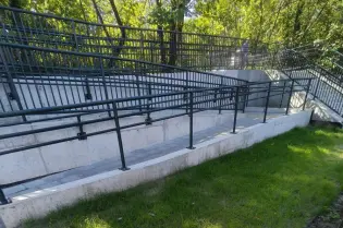 Widok na betonowe schody z barierkami otoczone zielenią.
