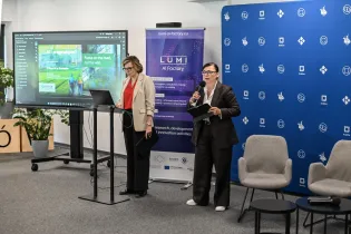 Kobieta przemawia do publiczności, obok druga osoba przy laptopie. W tle prezentacja na ekranie.