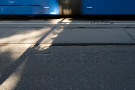 Niebieski tramwaj poruszający się po ulicy z widocznymi cieniami na jezdni.