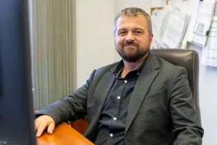 Mężczyzna w garniturze siedzący przy biurku, uśmiechający się do kamery.