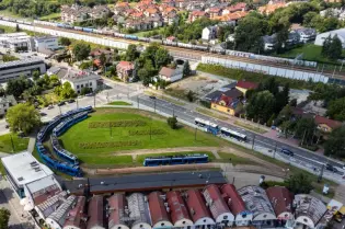 Tramwaj na pętli w otoczeniu zieleni i zabudowy miejskiej.