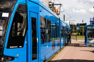 Niebieski tramwaj zatrzymany na przystanku, pasażerowie wsiadają i wysiadają.