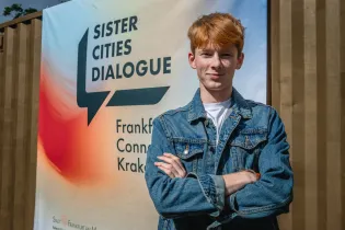 Młody mężczyzna w dżinsowej kurtce stoi przed banerem Sister Cities Dialogue.