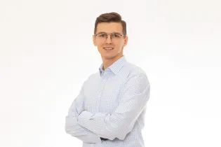 Mężczyzna w okularach, ubrany w niebieską koszulę w kratę, z uśmiechem i założonymi rękami.