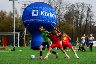 Dwie dziewczynki grają w piłkę nożną na boisku, w tle balon z logo Krakowa.