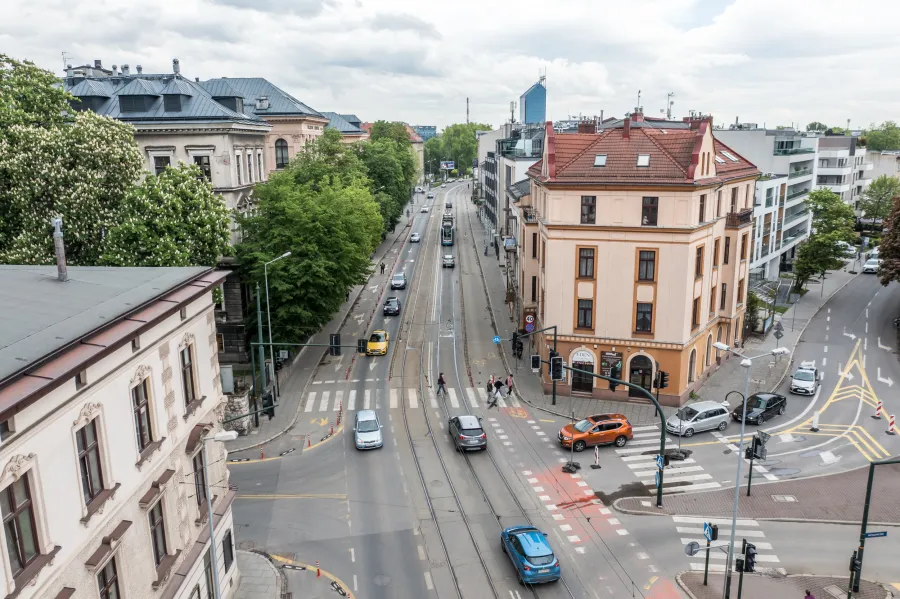 Widok na ruchliwą ulicę z tramwajami, samochodami i pieszymi, otoczoną zielenią i architekturą.