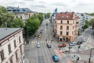 Widok na ruchliwą ulicę z tramwajami, samochodami i pieszymi, otoczoną zielenią i architekturą.