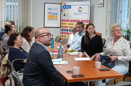 Zdjęcie przedstawia grupę osób siedzących przy stole podczas spotkania w sali Centrum Obywatelskiego Centrum C10. W tle widoczny jest baner Centrum oraz materiały informacyjne.