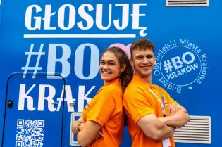 Dwie osoby w pomarańczowych koszulkach pozują z założonymi rękami na tle niebieskiej grafiki z napisem „GŁOSUJĘ #BO KRAKÓW” i kodem QR.