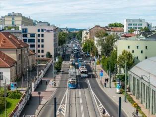 Widok na ruchliwą ulicę z tramwajami i samochodami, otoczoną budynkami i zielenią.