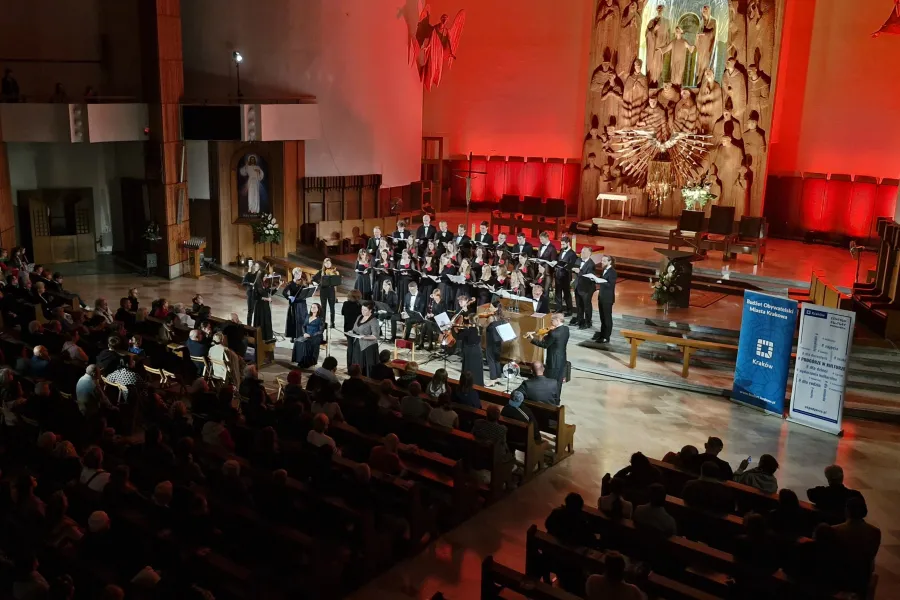 Koncert chóralny w kościele, oświetlony czerwonymi światłami, z publicznością siedzącą na ławkach.