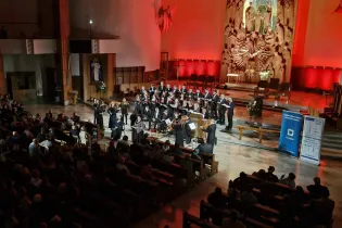 Koncert chóralny w kościele, oświetlony czerwonymi światłami, z publicznością siedzącą na ławkach.