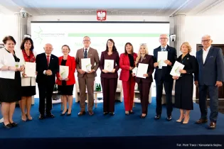 Grupa osób trzymających dokumenty, stojących na tle sali konferencyjnej.