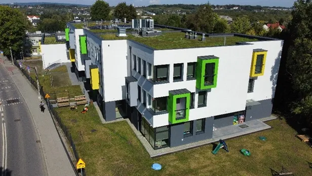 Nowoczesny budynek z zielonymi akcentami i zielonym dachem, otoczony trawnikiem.