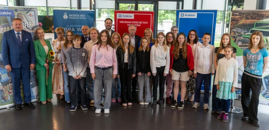 Grupa dzieci i dorosłych na wydarzeniu promującym Kraków, z banerami w tle.