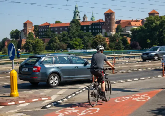 Rowerzysta na ścieżce rowerowej obok samochodu, w tle zamek na wzgórzu.
