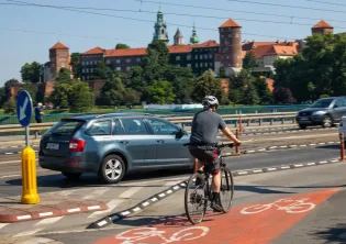 Rowerzysta na ścieżce rowerowej obok samochodu, w tle zamek na wzgórzu.
