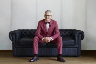 Mężczyzna w eleganckim garniturze siedzi na czarnej sofie w minimalistycznym wnętrzu.