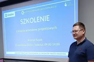 Zdjęcie przedstawia prowadzącego szkolenie stojącego obok ekranu z prezentacją „Szkolenie z pisania wniosków projektowych”. Na ekranie widoczne są także data i godziny szkolenia.