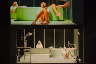 Scena teatralna z dwiema kobietami w białych szatach, jedna w wannie, druga na kanapie.