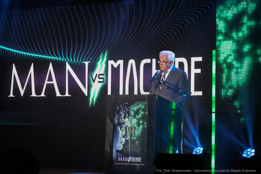 Na tle ledowewgo ekranu z napisem Man vs Machine ASPIRE 2026 przy mównicy na której wyświetla się graficzny motyw konferencji i antyczna głowa oraz cyfry przemawia Stanisław Kracik.