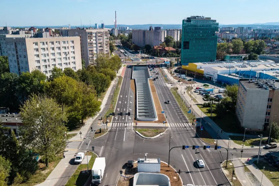 Widok na nowoczesną ulicę z rondem, otoczoną budynkami i zielenią.