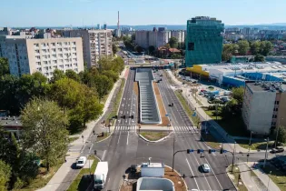 Widok na nowoczesną ulicę z rondem, otoczoną budynkami i zielenią.