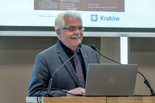Na podium, przy mównicy, na tle telebimu z widoczną częściowo prezentacją i logotypem Krakowa stoi uśmiechnięty siwy mężczyzna w czarnej koszuli z krawatem i popielatej marynarce który przemawia.    