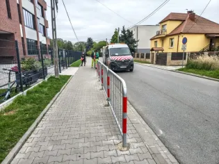 Przejście dla pieszych z barierkami, samochód dostawczy na ulicy, budynki w tle.