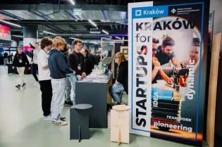 Stoiska promujące startupy w Krakowie, ludzie rozmawiają i wymieniają się informacjami.