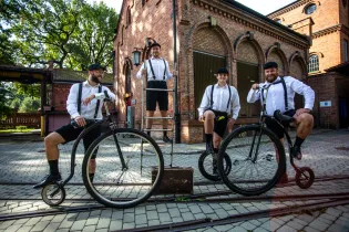 Czterech mężczyzn w strojach retro na rowerach, w tle zabytkowy budynek.