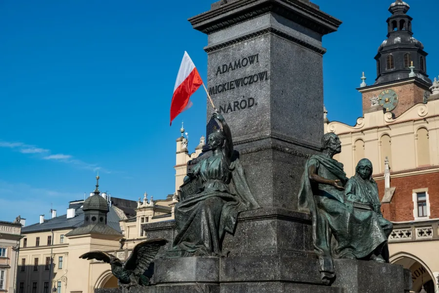 Pomnik Adama Mickiewicza z flagą Polski w tle, otoczony architekturą Krakowa.