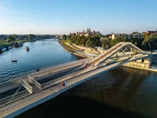 Nowoczesny most nad rzeką z widokiem na zieleń i zabudowę miejską.
