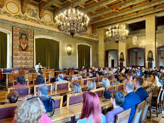 Sala konferencyjna z dużą liczbą uczestników, prelegent przy mównicy, eleganckie wnętrze.