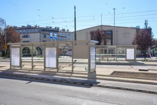 Pusta wiata przystankowa z reklamami w trakcie wymiany, w tle budynki mieszkalne.