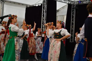 Grupa dziewcząt w tradycyjnych strojach ludowych tańczy i śpiewa na scenie.