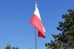 Grafika przedstawia flagę Polski na tle niebieskiego nieba.