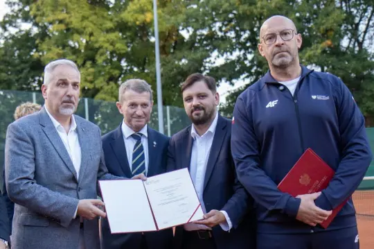 Czterech mężczyzn trzyma dokumenty, w tle kort tenisowy i drzewa.