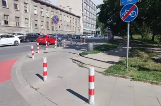 Ulica z oznaczeniami drogowymi, słupkami i parkowanymi samochodami.