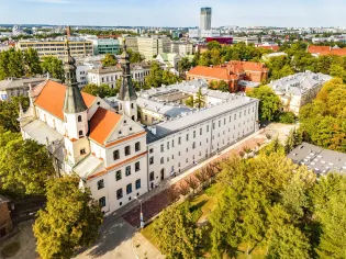 Widok na zabytkowy budynek z wieżami i otaczającą go zieleń.