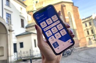Ręka trzymająca telefon komórkowy z otwartą aplikacją mKraków. W tle widoczny kościół Franciszkanów przy ulicy Franciszkańskiej w Krakowie.