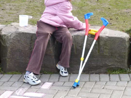 na szarym murku siedzi dziecko w brązowych spodniach i różowej zimowej kurtce z kapturem. Nie widzimy jego twarzy. Obok stoją oparte na murku kolorowe dziecięce kule inwalidzkie.  