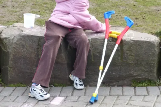 Dziecko w różowej kurtce siedzi na kamieniu, obok leżą kule ortopedyczne.