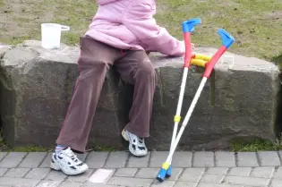 Dziecko w różowej kurtce siedzi na kamieniu, obok leżą kule ortopedyczne.