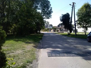 Droga w kierunku zabudowań, otoczona drzewami i zielenią.