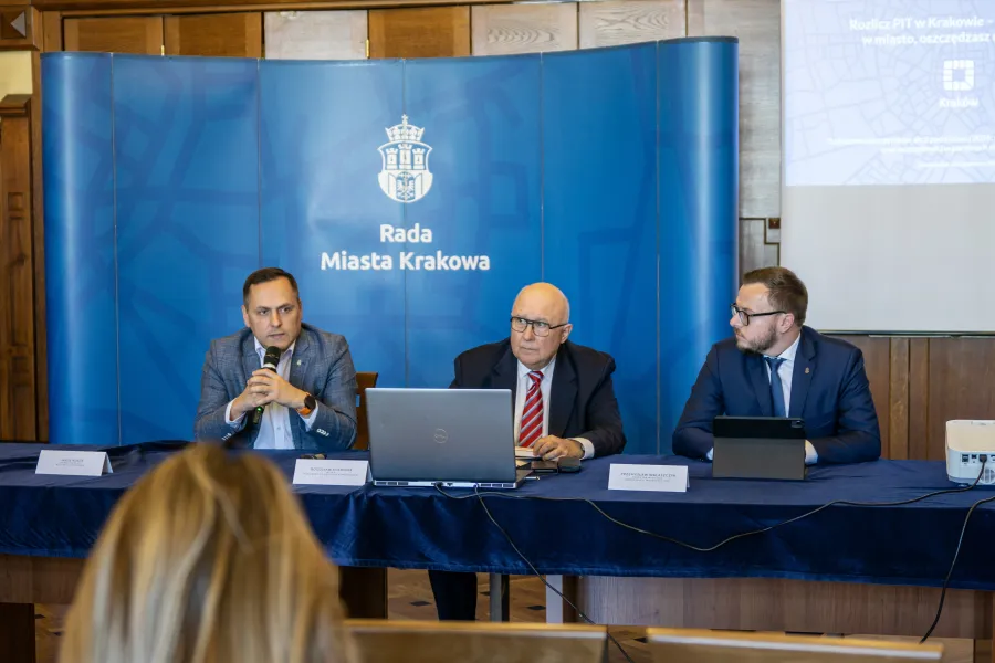 Konferencja Rady Miasta Krakowa z trzema mężczyznami przy stole i prezentacją na ekranie.