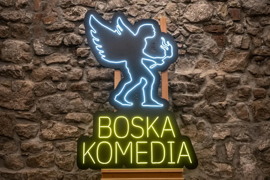 Neonowy znak z napisem Boska Komedia i postacią anioła trzymającego pochodnię.