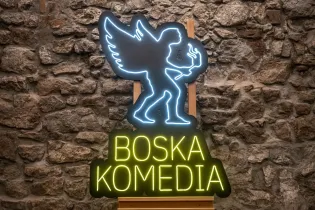 Neonowy znak z napisem Boska Komedia i postacią anioła trzymającego pochodnię.